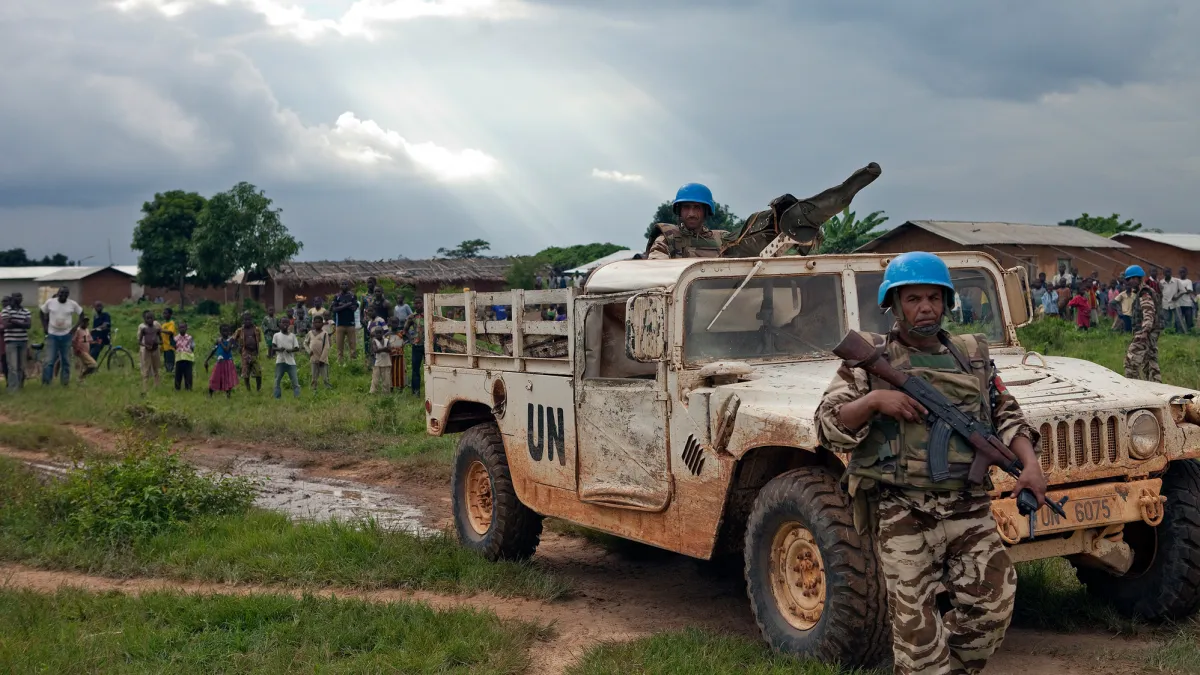 Two peacekeepers in uniform wearing blue helmets and holding weapons. One of the peacekeepers is standing in a truck which says 'UN" on the side.