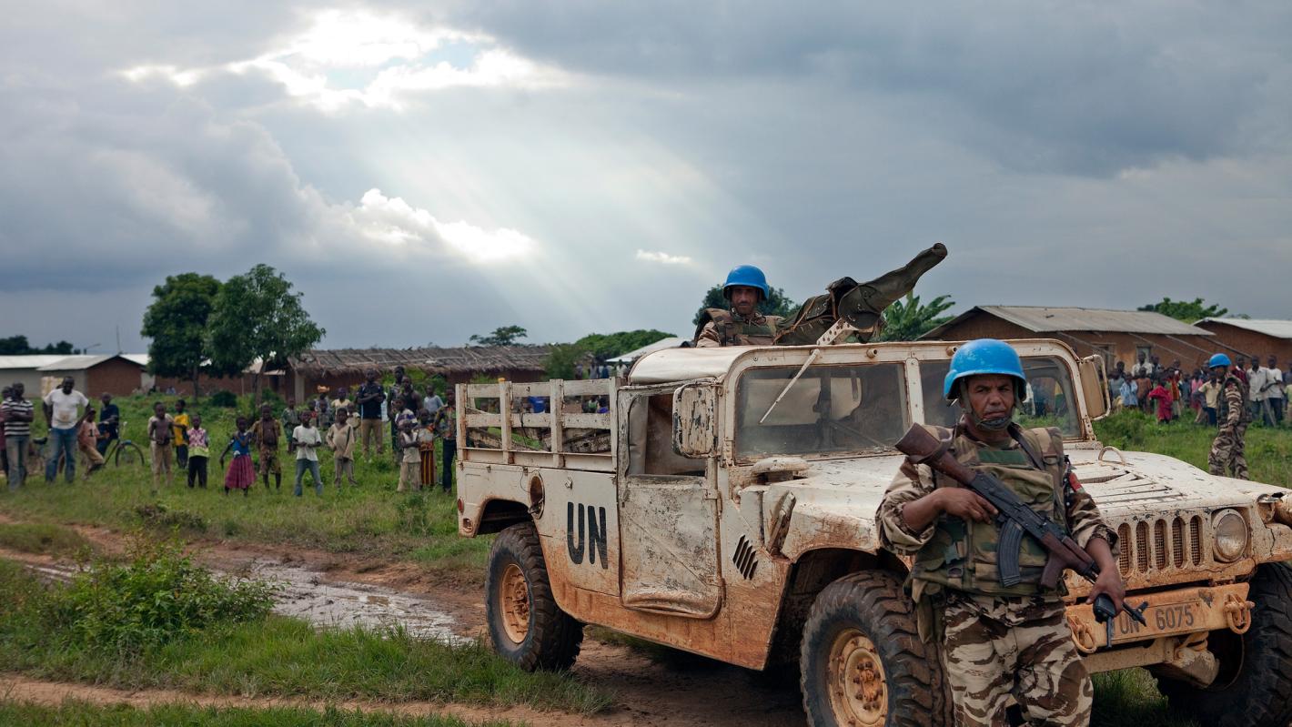 Two peacekeepers in uniform wearing blue helmets and holding weapons. One of the peacekeepers is standing in a truck which says 'UN" on the side.