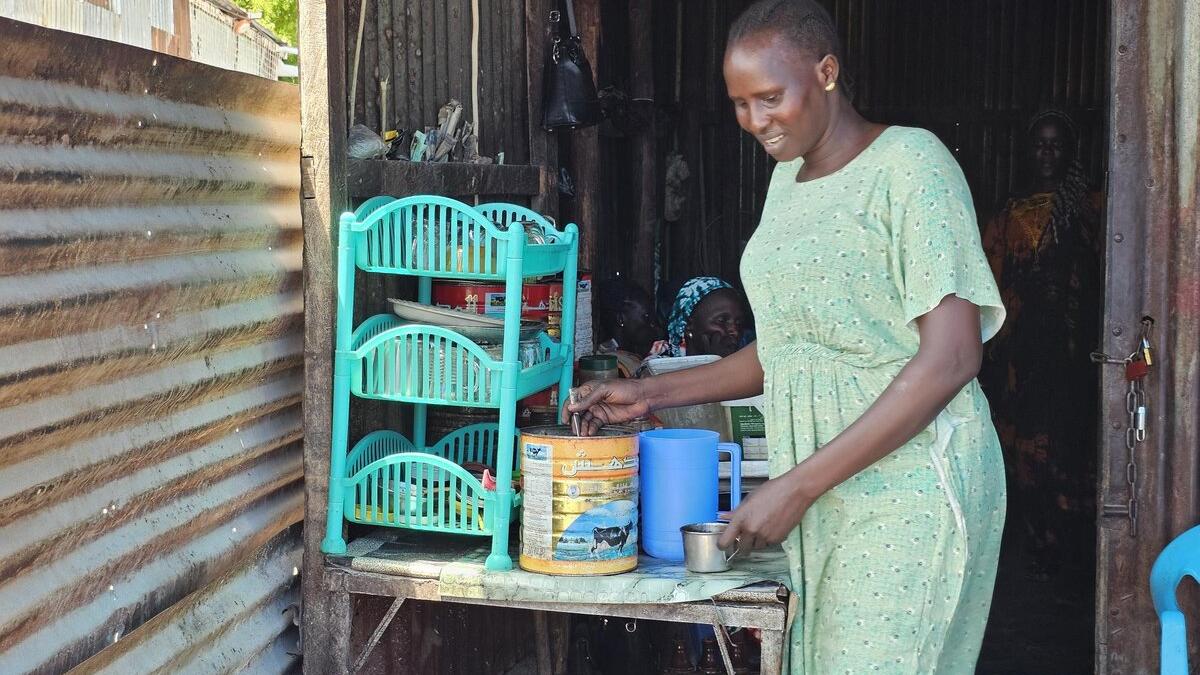 A person in a light green dress stands at a small table inside a corrugated metal structure, preparing items near plastic shelves and containers.