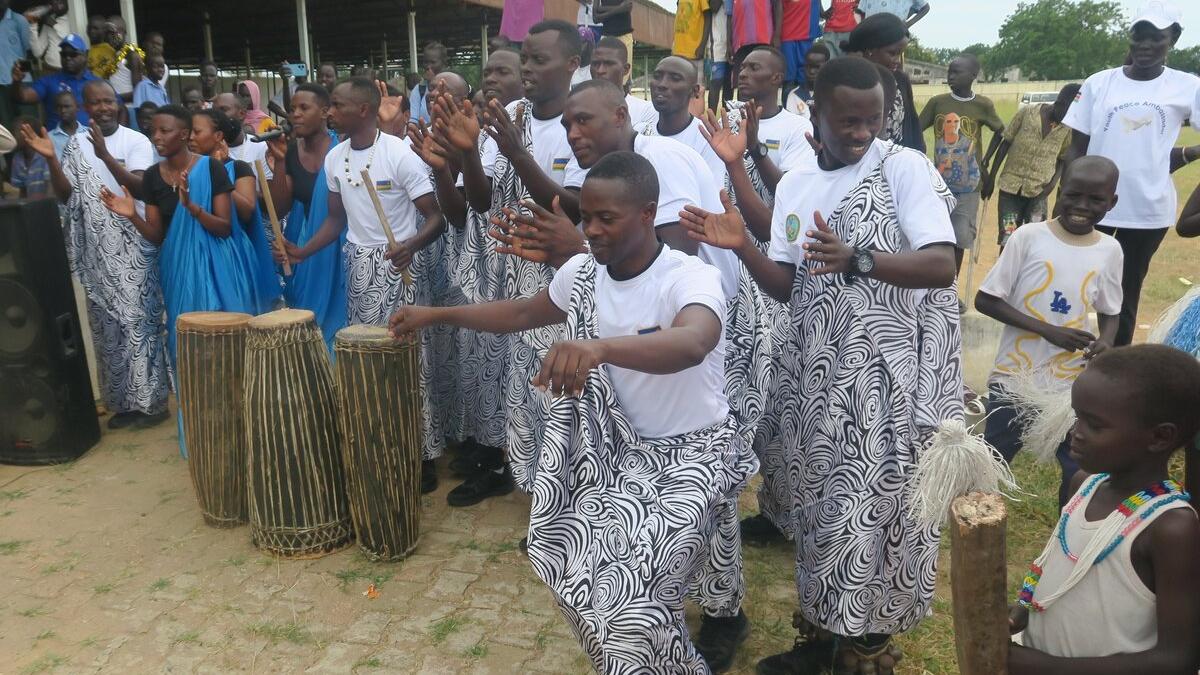 Group of people wearing white shirts and patterned wraps performing a traditional dance outdoors, with drums placed in front and others clapping along.