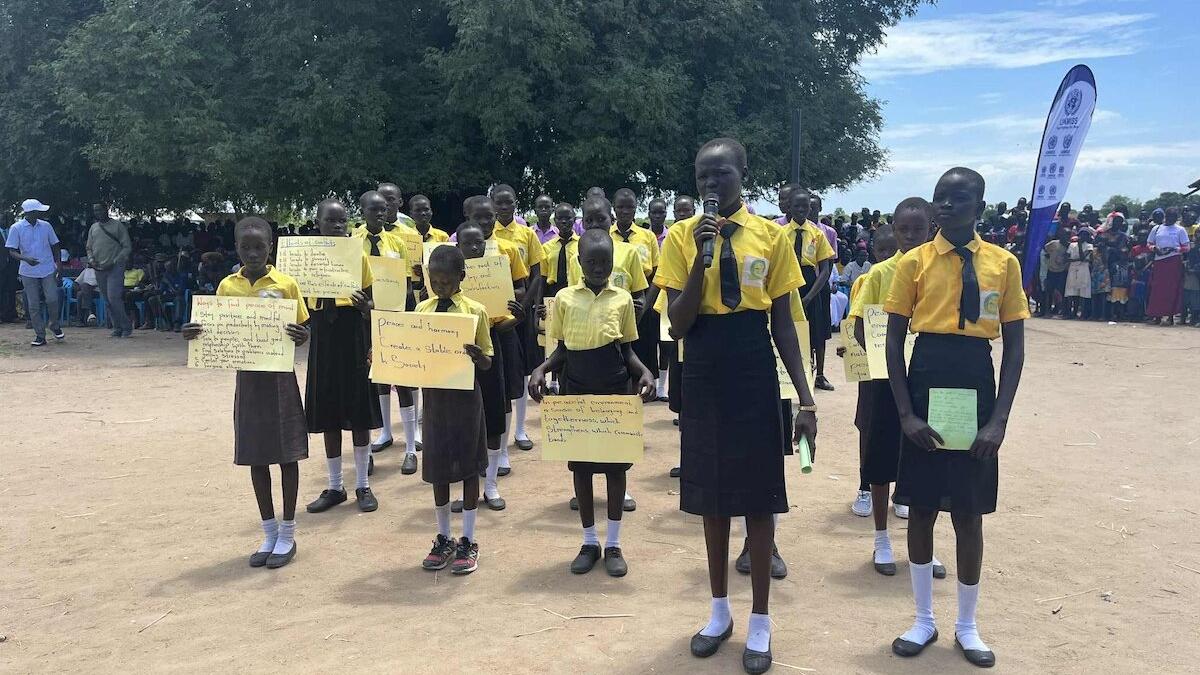 Group of schoolchildren in yellow shirts and dark skirts or trousers holding handwritten signs and papers during an outdoor event, with a crowd and banner in the background.
