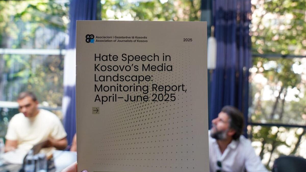 A close-up of a hand holding a report titled “Hate Speech in Kosovo’s Media Landscape: Monitoring Report, April–June 2025” published by the Association of Journalists of Kosovo. The background shows a meeting setting with people seated at a table and large windows with greenery outside.