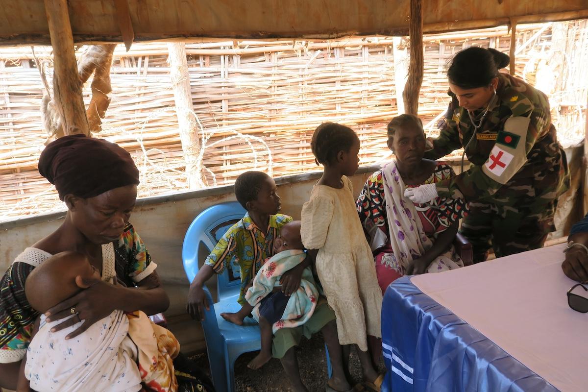 A uniformed medical officer is providing treatment to a seated woman. Next to the female patient is another woman and three children. 