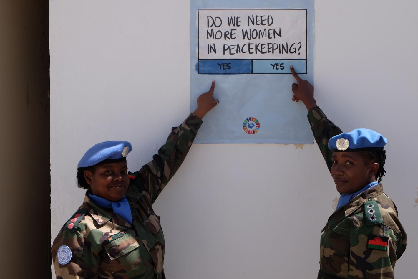 Do we need more women in peacekeeping? Yes or yes? Two women peacekeepers in military uniform point at a sign that poses the question "do we need more women in peacekeeping" and gives "yes" or "yes" as possible responses.