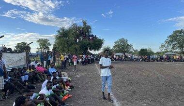 Large crowd gathered around a dirt sports field, with people sitting along the sidelines and others perched in a tree to watch the event.