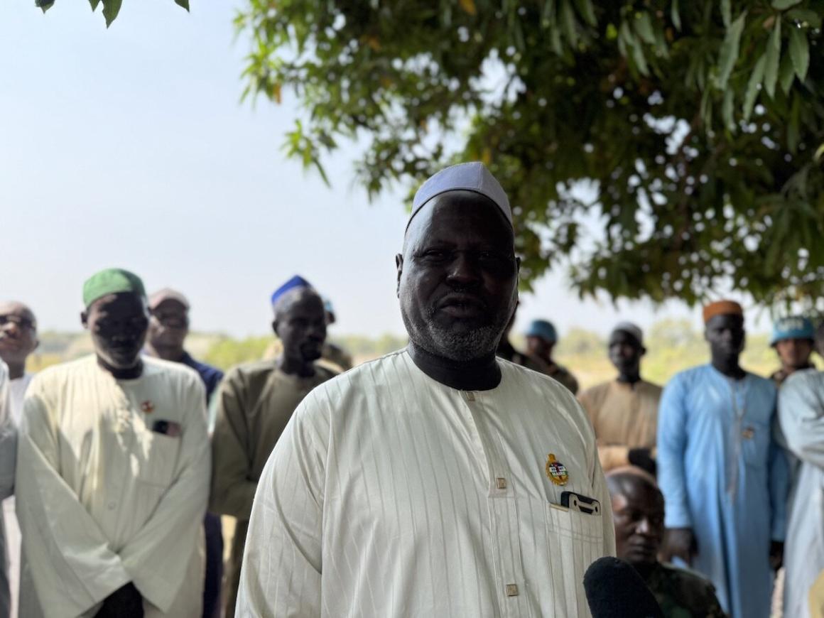 A man stands in the foreground with a group of men bhind him, under a tree