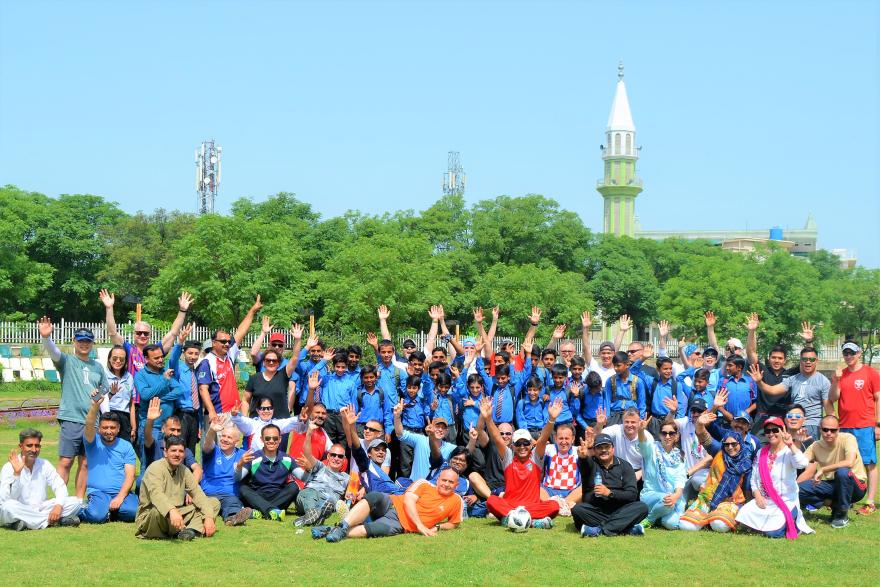 Large group of people sitting and standing on grass, raising hands for a group photo in front of trees and a tall white tower.